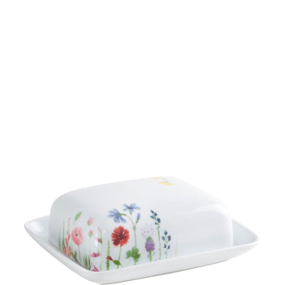 Eine weiße Butterdose mit farbenfrohen, blühenden Blumenmustern auf der Vorderseite. Die Dose hat eine rechteckige Form und einen leicht gewölbten Deckel. Das florale Design umfasst verschiedene Blumen wie Mohn und Kornblumen, die fröhlich und bunt wirken. Die Dose steht auf einer leicht geschwungenen Unterlage und hat eine glatte Oberflächenstruktur.