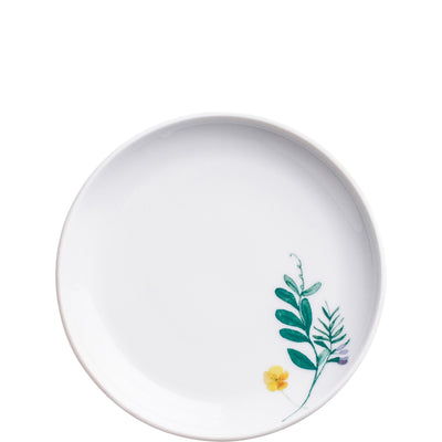 Ein runder, flacher Porzellanteller mit einem weißen Hintergrund. Am Rand des Tellers befindet sich eine stilisierte Darstellung von grünen Blättern und einer kleinen gelben Blüte sowie einem lila Akzent, die ihm ein leicht florales Design verleihen. Der Teller wirkt elegant und modern.