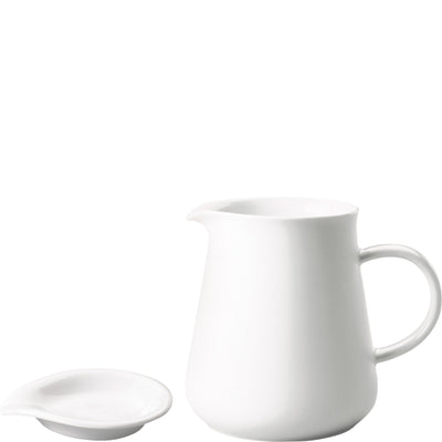 Ein schlichtes, weißes Kännchen aus Porzellan steht neben einer flachen Untertasse, ebenfalls in Weiß. Das Kännchen hat eine elegante, geschwungene Form mit einem leicht ausgeformten Ausguss und einem angenehmen Henkel. Die Untertasse hat eine runde Form und ist für die Ablage des Kännchens geeignet. Beide Teile sind minimalistisch gestaltet und wirken modern und zeitlos.