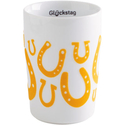 Weißer Keramikkbecher mit bunten gelben Hufeisen-Motiven, die um den Becher herum angeordnet sind. Auf der Oberkante des Bechers steht in schwarzer Schrift das Wort "Glückstag". Der Becher hat eine schlichte, moderne Form und vermittelt eine fröhliche und einladende Atmosphäre. Ideal für Kaffeeliebhaber oder als Geschenk.