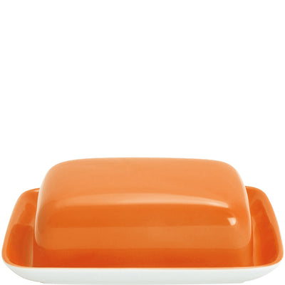 Eine orangefarbene Butterdose ist auf einem weißen Unterteller platziert. Die Dose hat eine glänzende Oberfläche und eine rechteckige Form mit abgerundeten Kanten. Der Unterteller ist ebenfalls rechteckig und hat eine schlichte, weiße Ausführung. Das Bild zeigt die Butterdose von oben.