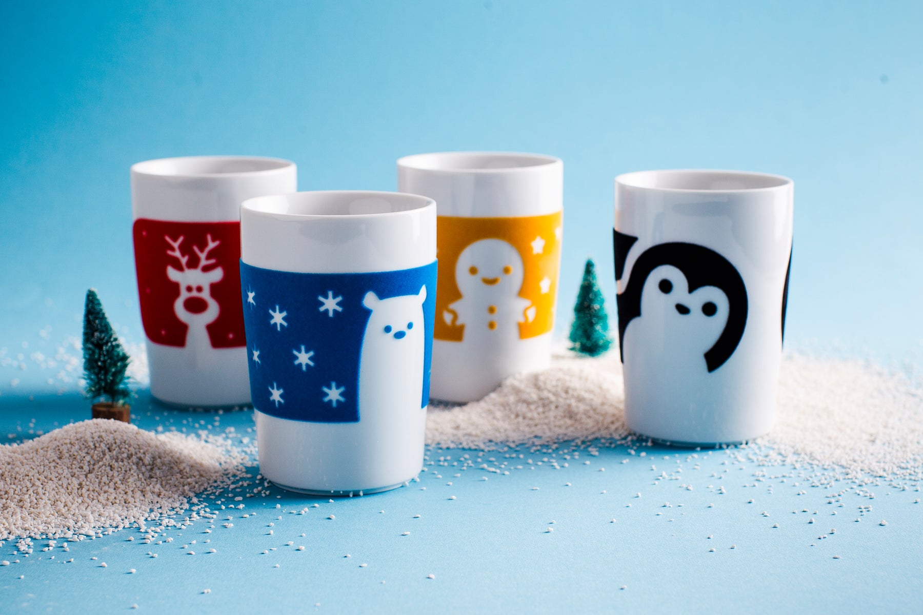 Vier Tassen mit winterlichen Motiven stehen auf einem blauen Hintergrund. Jede Tasse zeigt unterschiedliche Designs: eine mit einem Rentier, eine mit einem Schneemann, eine mit einem niedlichen Eisbären auf blauer Basis und eine mit einem Pinguin. Um die Tassen herum liegen kleine, künstliche Weihnachtsbäume und eine Schicht aus weißem Material, das an Schnee erinnert. Die Farben der Tassen sind rot, blau, gelb und schwarz, was eine festliche Stimmung vermittelt.